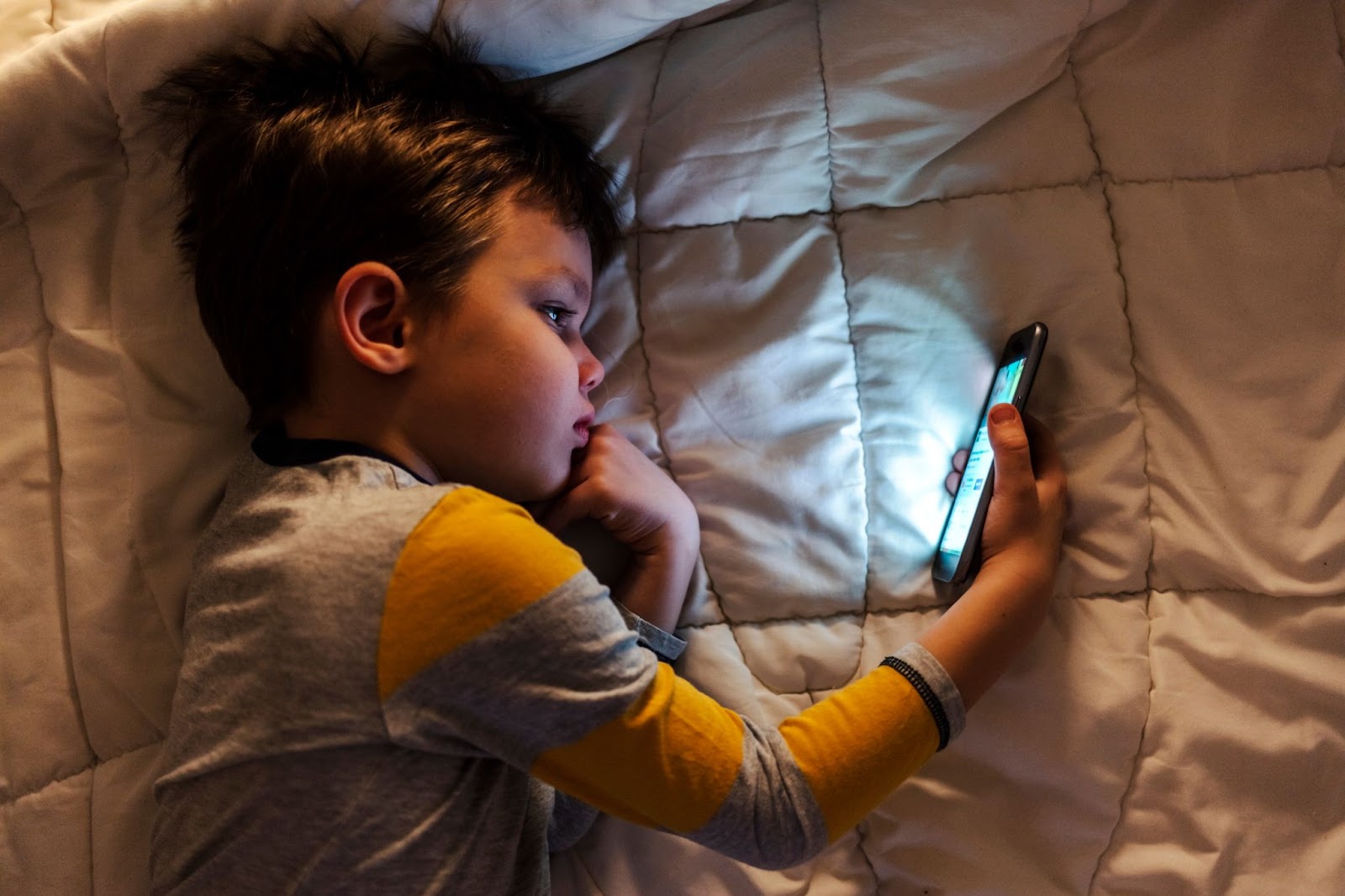 Gülümseyen yürümeye başlayan çocuk yatakta uzanıyor ve akıllı telefonuna bakıyor.